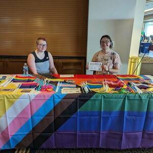 Two smiling students sit behind a table covered with the progress pride flag. The table features a variety of mini pride flags and stickers.