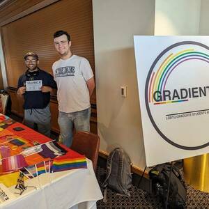 Two smiling students stand behind a table covered in pride flags and stickers, next to a large sign with the GradientND logo