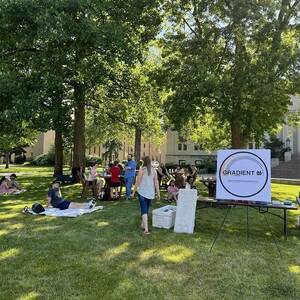 Wide shot of several picnic tables outside with several people sitting, standing, and talking and a large GradientND sign in the foreground.