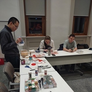 Photo of tables with various cookie-decorating supplies. Three students are seated or standing nearby, decorating cookies.