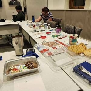 Photo of tables with cookies and decorating supplies, with two students on the far side of the table decorating cookies. There is a container of decorated cookies in the foreground.