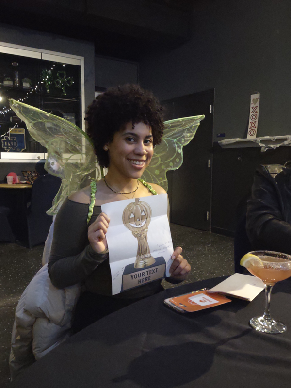 Photo of a black woman dressed as a fairy, smiling and holding up a printout of a Halloween-themed trophy.